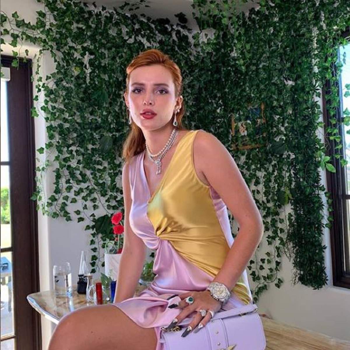 Bella Thorne poarta o rochie aurie cu violet, are parul prins si sta pe o masa