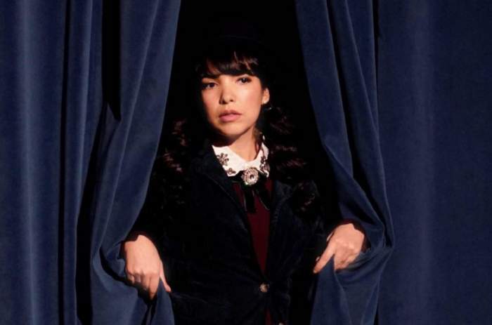 Indila se află în spatele unei cortine de teatru. Artista poartă un joben pe cap.