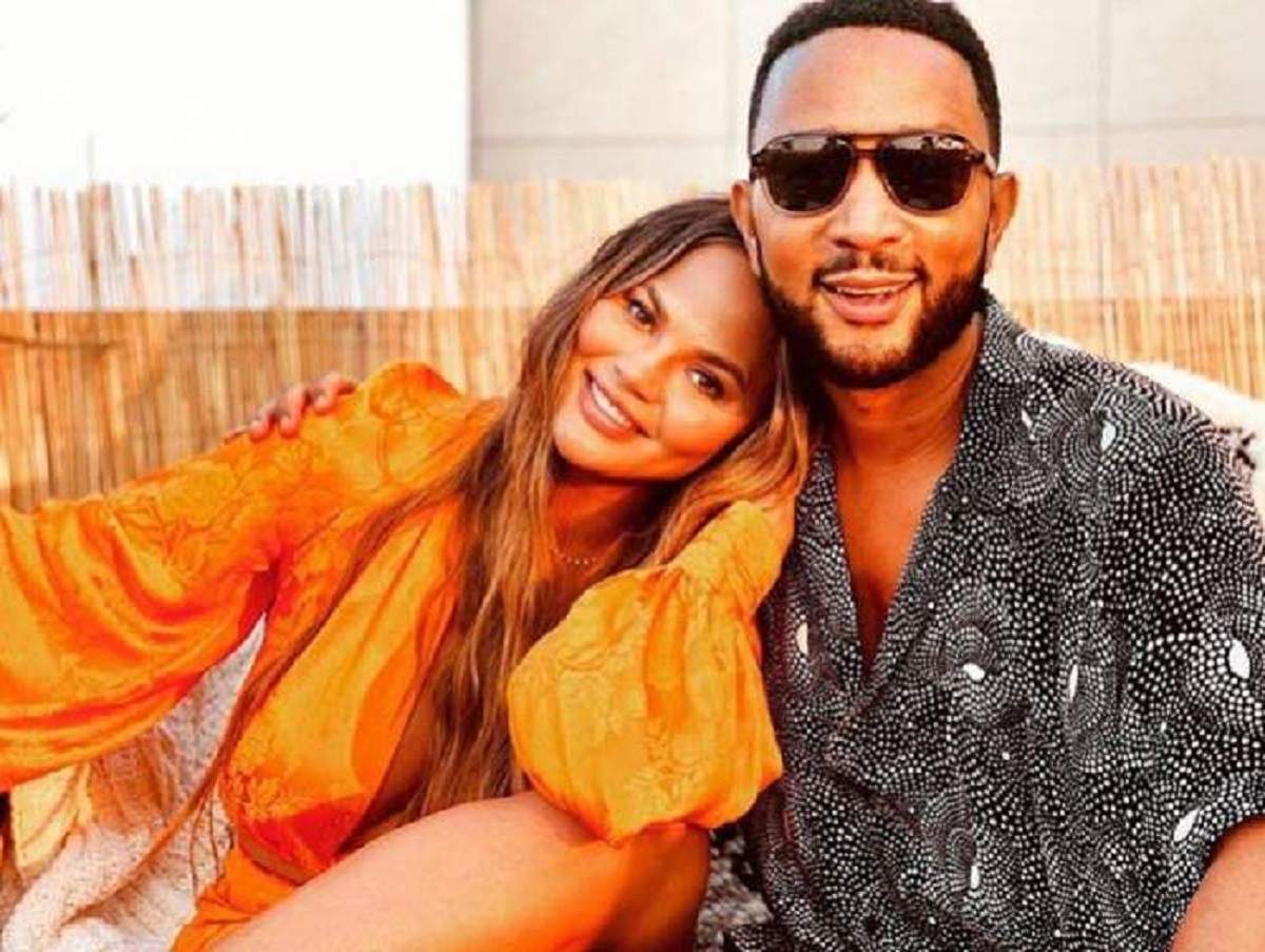 Chrissy Teigen si John Legend zambesc fericiti intr-o fotografie, ea poarta cu rochie, portocalie si el o camasa neagra si ochelari de soare