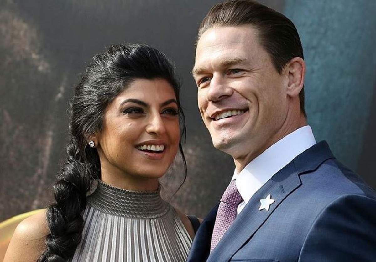 John Cena și Shay Shariatzadeh se află pe covorul roșu. Ea poartă o rochie gri și el un costum albastru.