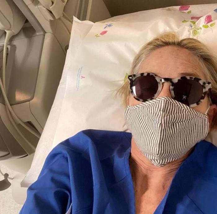 Sharone Stone e pe patul de spital, are masca pe fata si ochelari de soare