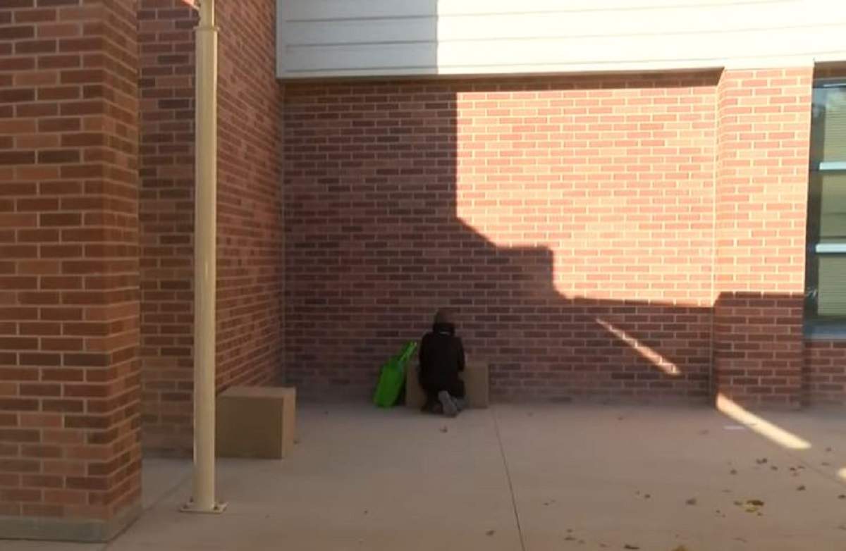 Un băiețel de 9 ani stă pe jos și își face temele pe trotuarul de lângă școală. Laptopul lui este așezat pe o cutie de carton.