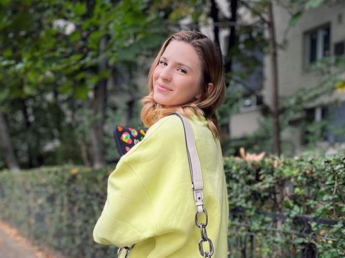 Cristina Ciobănașu poartă o bluză galbenă. Artista are pe umăr o geantă și zâmbește discret.
