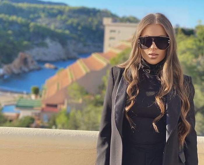 Oana Radu se află în vacanță. Artista poartă un sacou negru și ochelari de soare. În spatele ei se văd munții și marea.
