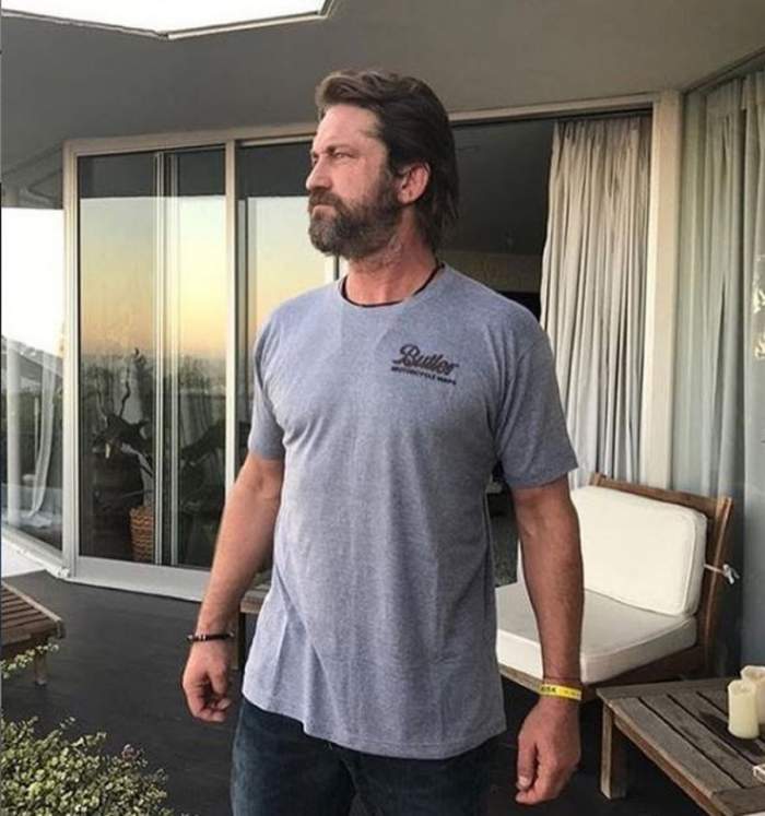 Gerard Butler sta pe terasa casei, poarta un tricou gri si se uita in drepta