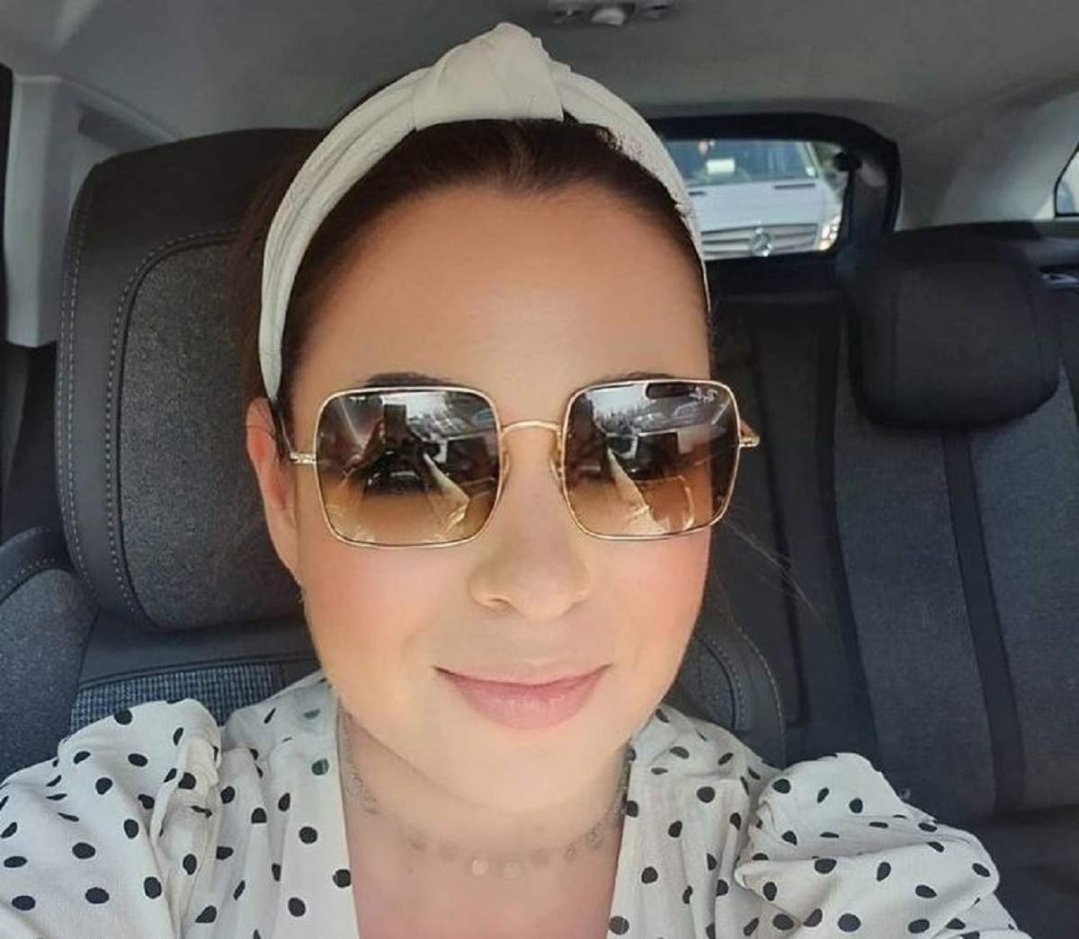 Oana Roman se află în mașină. Vedeta poartă o pereche de ochelari de soare și e îmbrăcată cu o cămașă albă, cu buline negre.