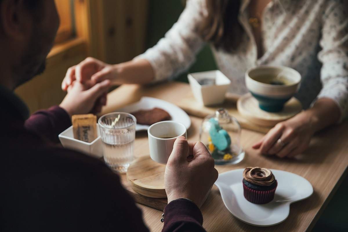 O femeie și un bărbat se ține de mână, în timp ce stau la o masă, la cafenea. Aceștia au în față prăjituri și cești de cafea.