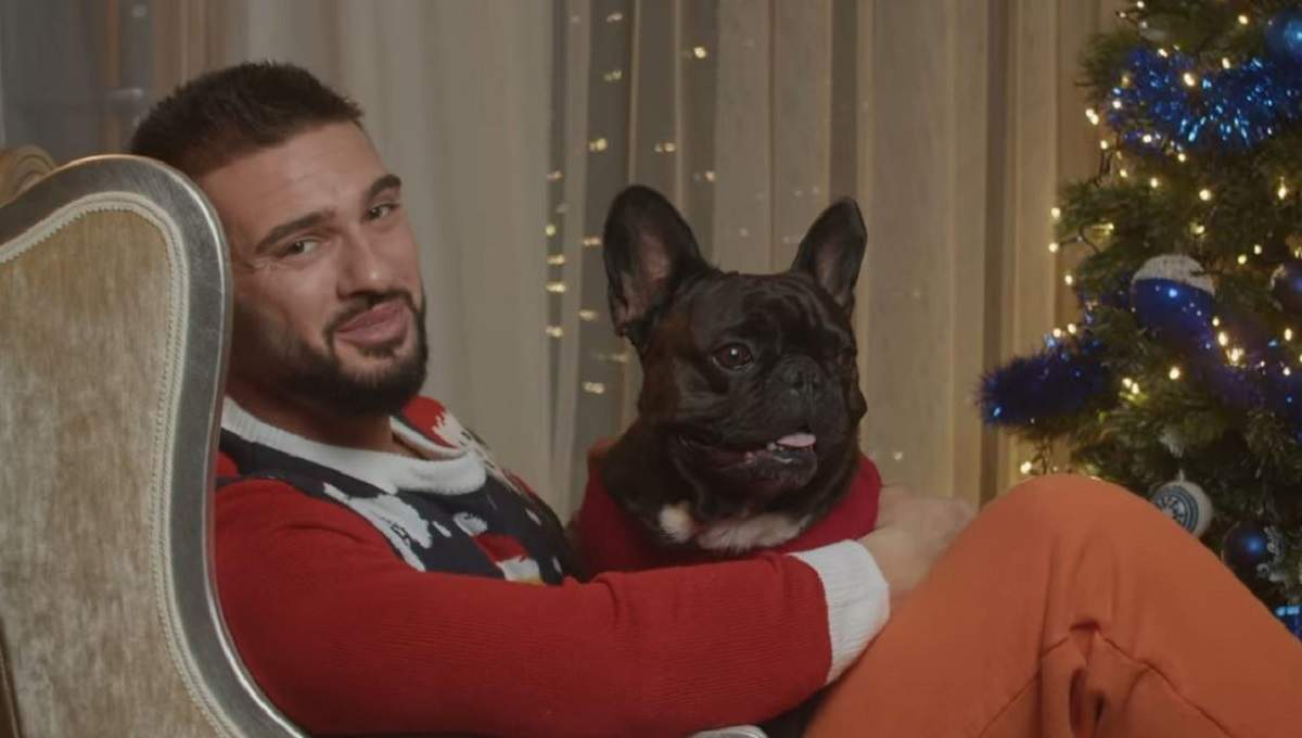 Dorian Popa este îmbrăcat cu un pulover roșu. Artistul stă pe fotoliu și îl ține în brațe pe câinele lui, Cheluțu. Lângă ei se vede un brad de Crăciun.