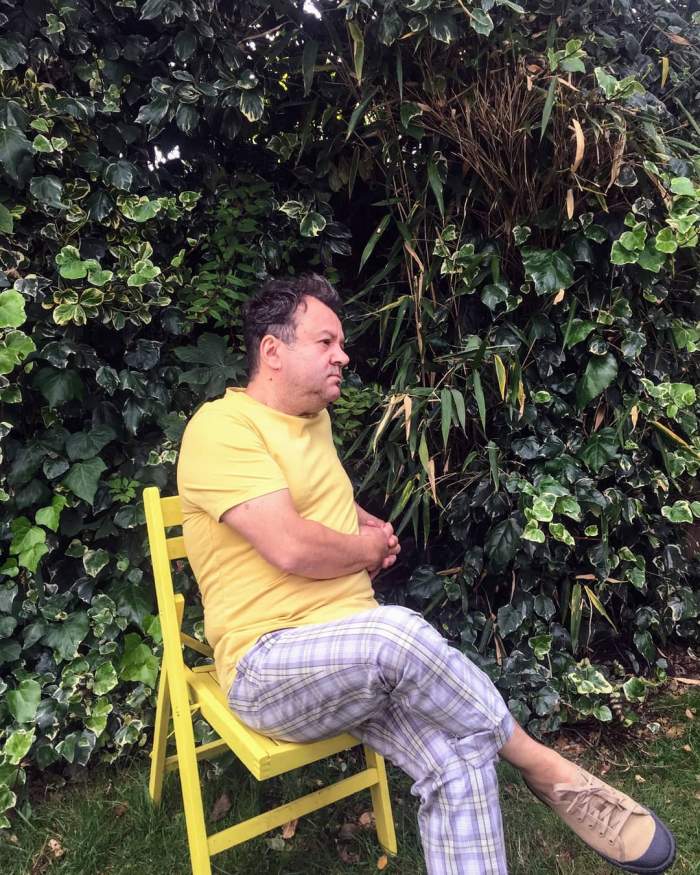 Dragoș Moștenescu s-a fotografiat stând pe un scaun galben, în grădină. în tricou galben și pantaloni albaștri