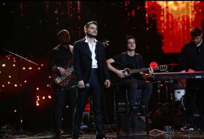 Adrian Petrache este pe scena in finala X Factor, poarta un costum negru si camasa alba descheiata la primii nasturi