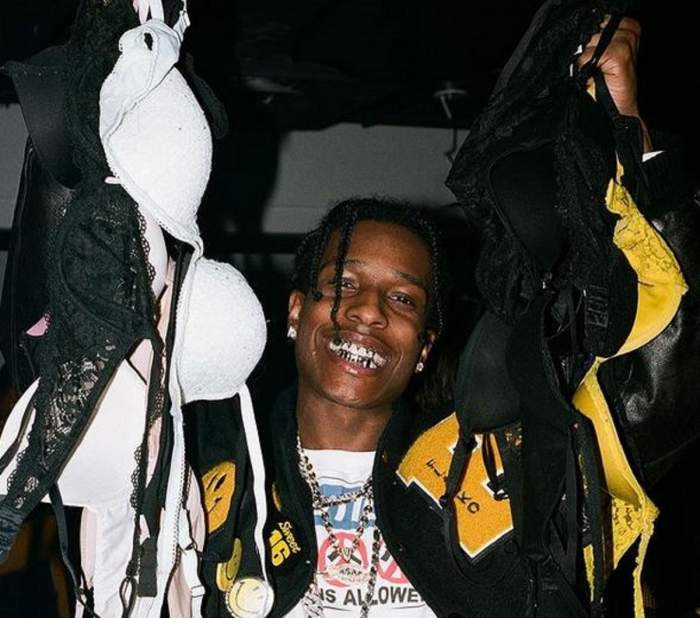 Asap Rocky poartă un tricou alb. Rapper-ul ține în mâini mai multe sutiene colorate și zâmbește larg.