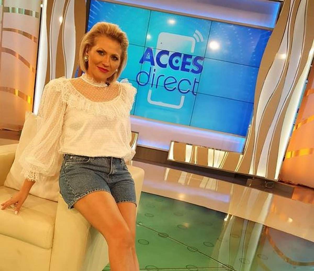 Mirela Vaida se află în platoul „Acces Direct”. Vedeta poartă o bluză albă și niște pantaloni scurți din denim și stă pe scaun.