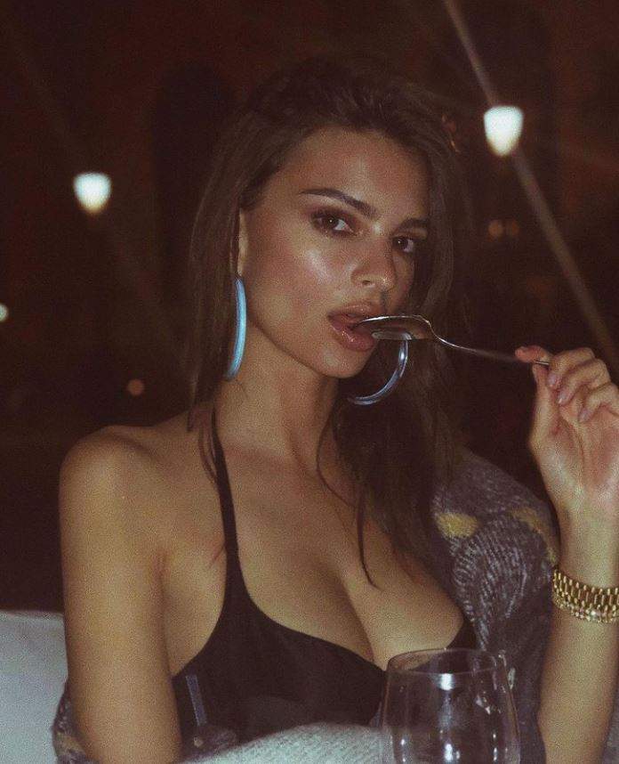 Emily Ratajkowski este la masa la restaurant, poarta un top decoltat si tine furculita intre dinti