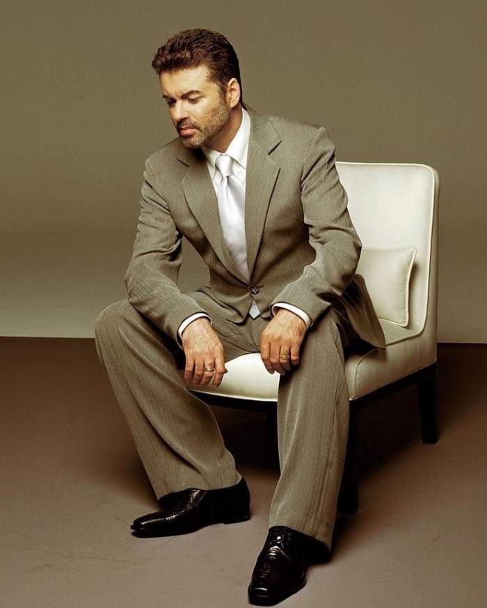George Michael, îmbrăcat în costum, stând pe un fotoliu