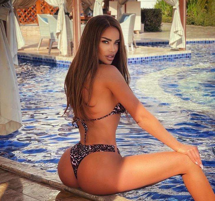 Bianca Drăgușanu stă așezată pe marginea piscinei. Vedeta își ține mâinile pe genunchi și poartă un costum de baie maro, cu pete albe.