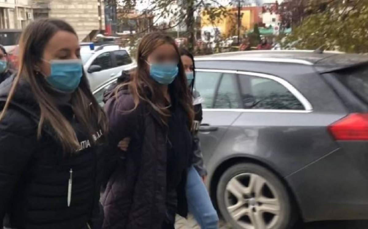 Soția geloasă din Iași care a răpit-o, bătut-o și tuns-o pe amanta soțului ei este dusă de două polițiste. Femeia poartă o geacă lungă, neagră.