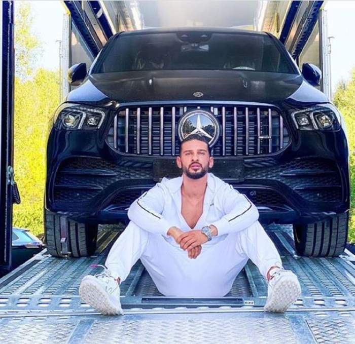 Dorian Popa stă în fața noii sale mașini, de culoare neagră, care se află încă pe platformă, fiind îmbărcat cu o ținută albă