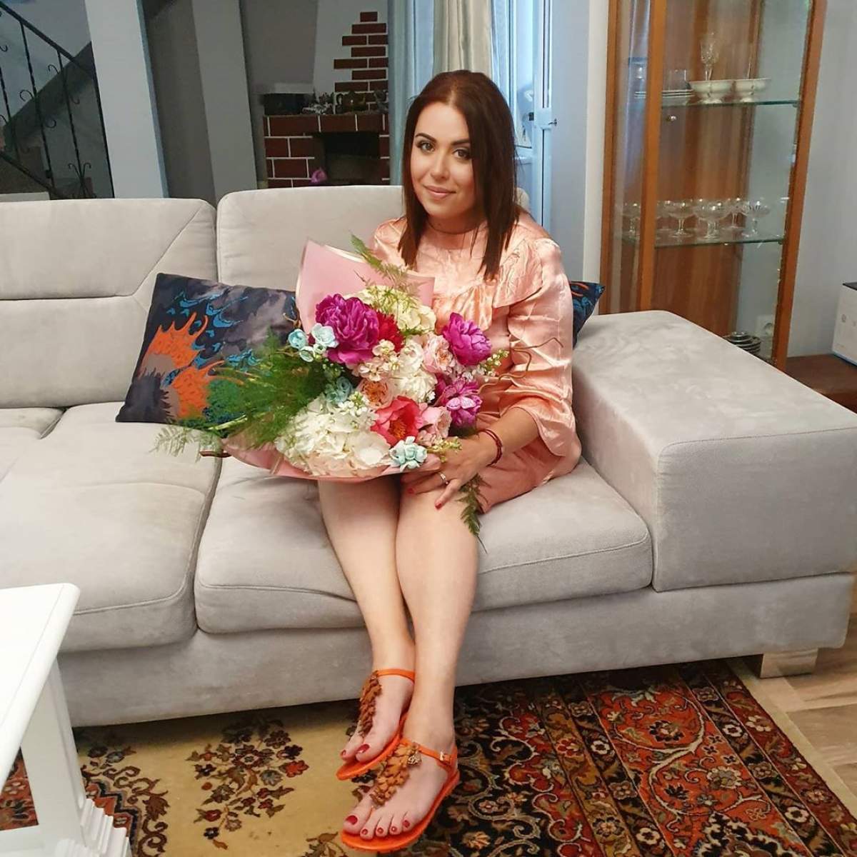 Oana Roman, îmbrăcată într-o rochie, stă pe canapea în timp ce ține un buchet de flori în brațe