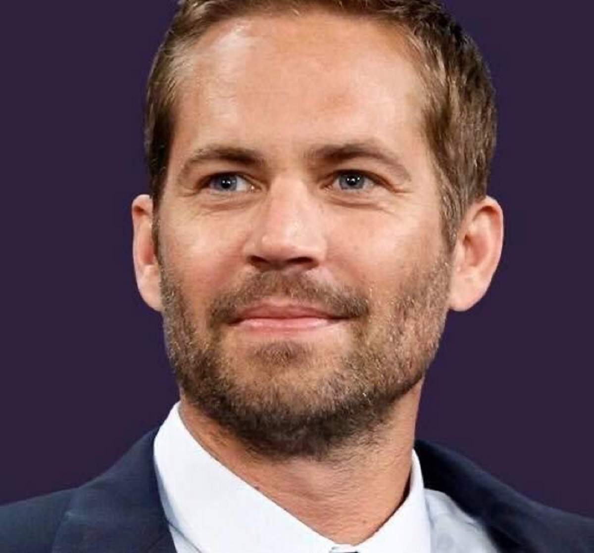 Paul Walker poartă un costum albastru închis și o cămașă albă. Actorul zâmbește și se uită într-o parte.