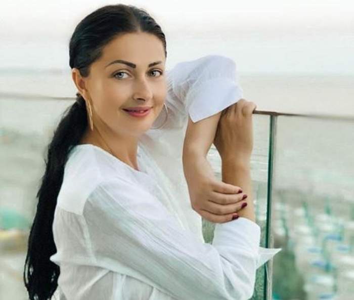 Gabriela Cristea e pe balcon. Vedeta are brațul pe balustradă și e îmbrăcată într-o cămașă albă.
