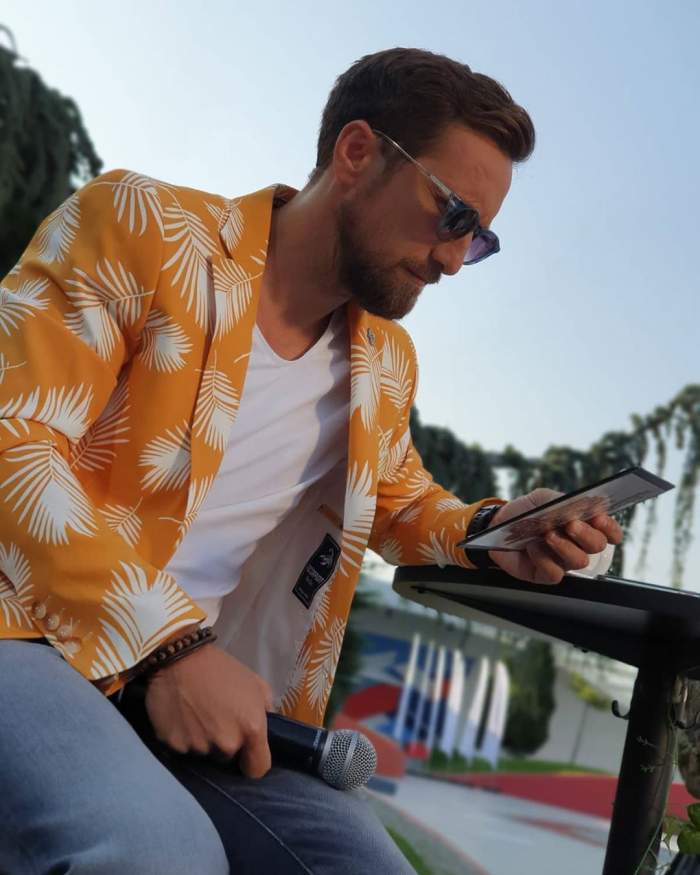 Dani Oțil butonează telefonul, purtând un sacou portacaliu, în timpul emisiunii pe care o prezintă