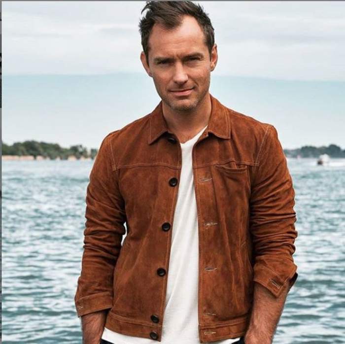 Jude Law are în spate un lac, poartă un tricou alb și jachetă maro