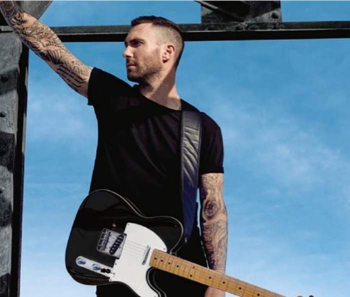 Adam Levine se spijină de un stâlp, are chitara agățată de umăr și se uită în partea dreaptă