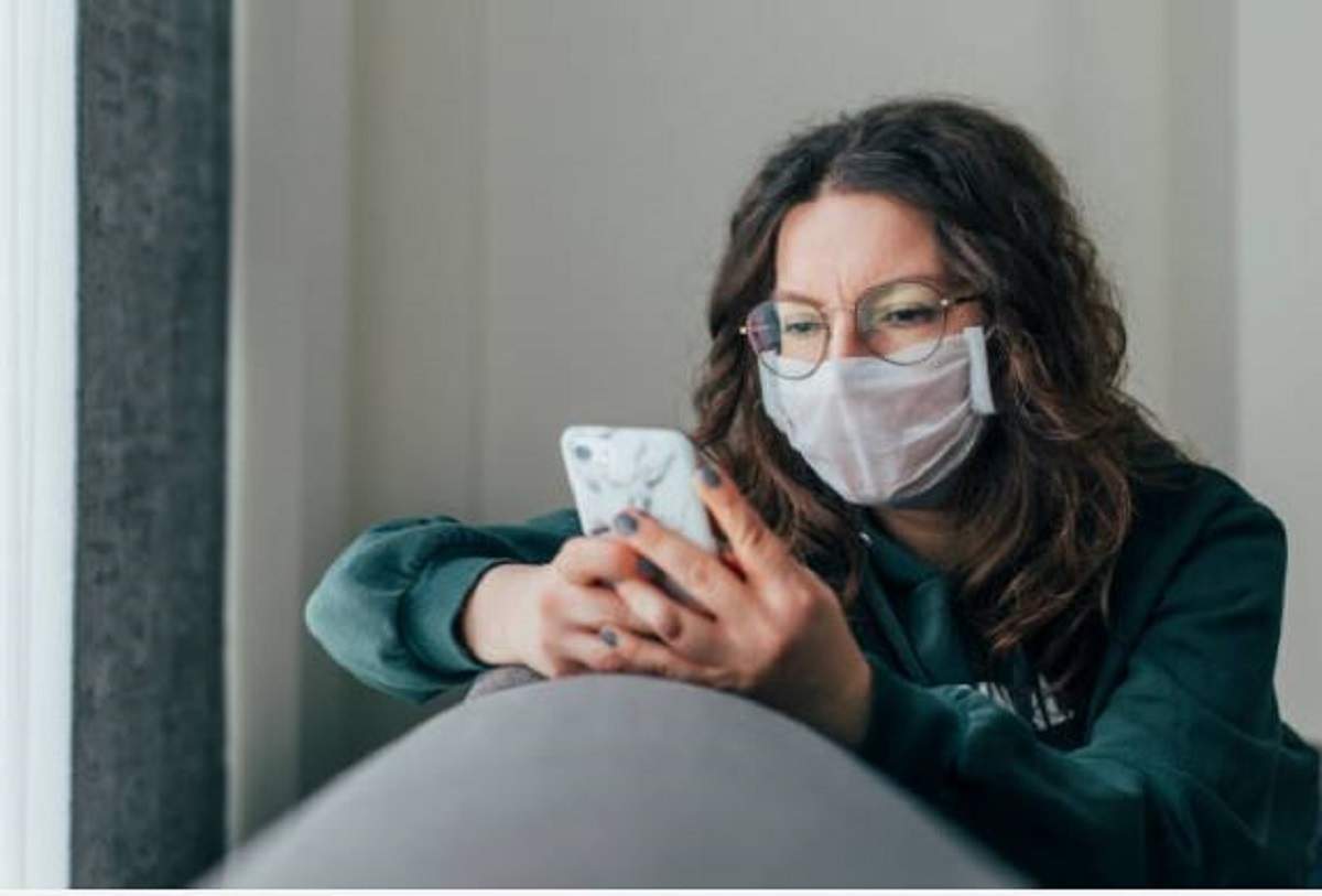 Imagine ilustrativa cu o femeie care poarta ochelari si masca de protectie pe fata, in timp ce se uita pe telefon