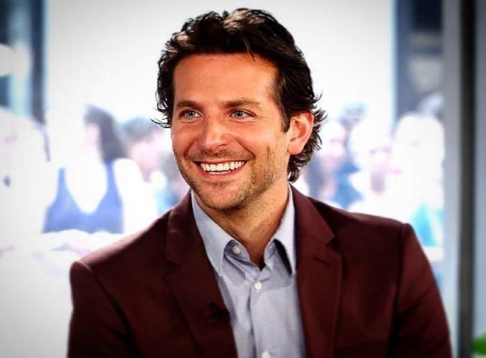 Bradley Cooper zâmbește larg, în timp ce poartă un sacou maro și o cămașă albastră
