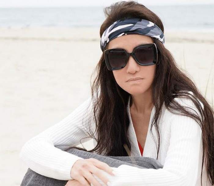 Vera Wang stă pe nisip, la mare. Vedeta poartă o pereche de ochelari de soare și o bluză albă