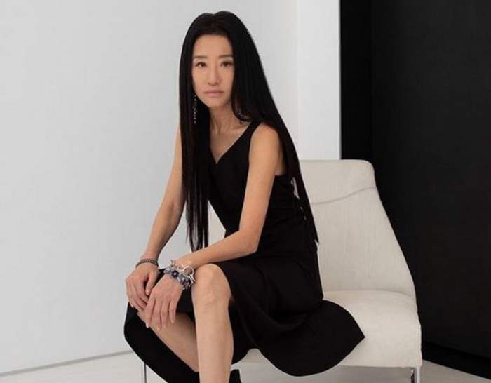 Vera Wang stă pe un scaun alb. Creatoare de modă poartă o rochie neagră, lungă