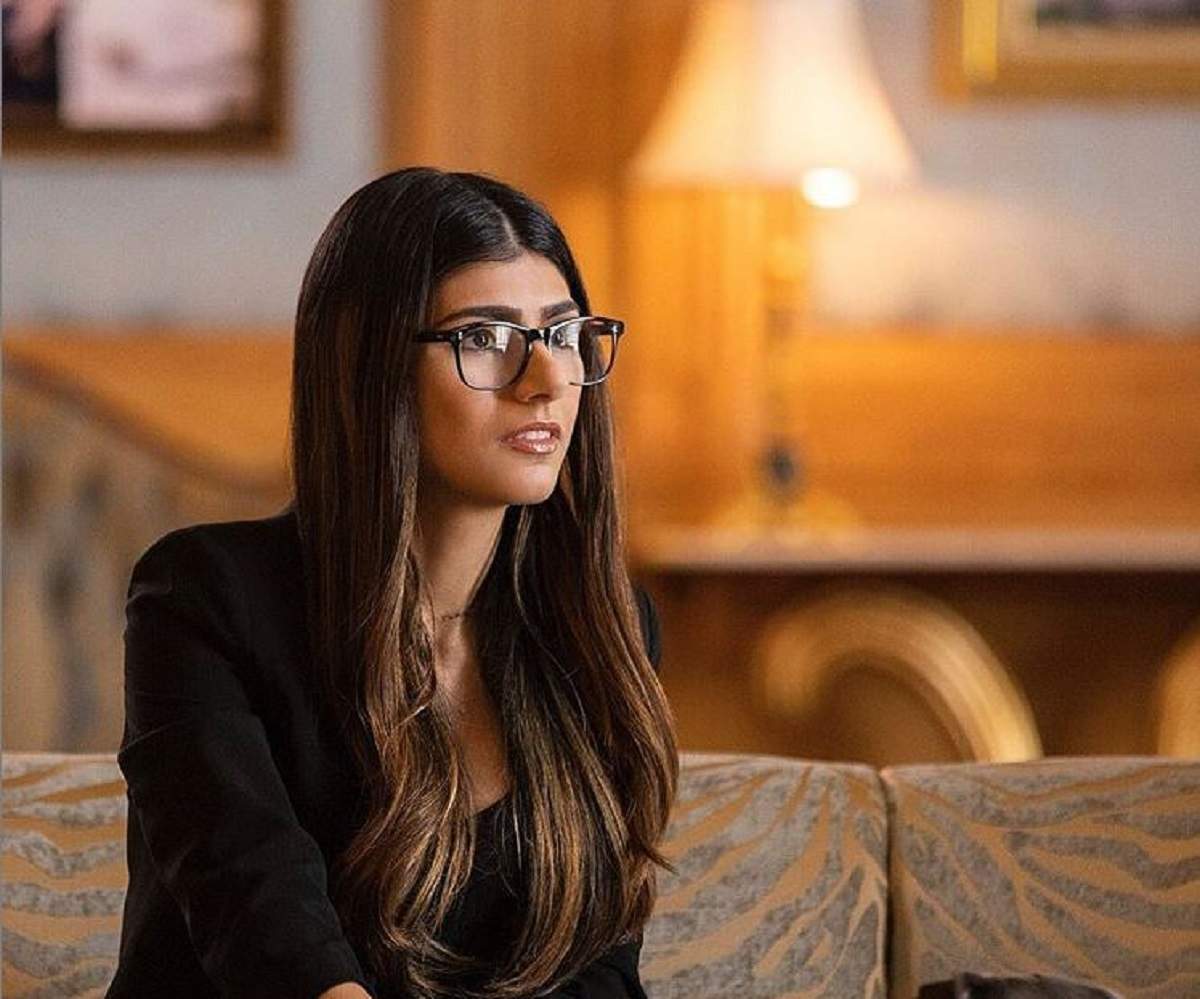 Mia Khalifa este imbracata cu un sacou negru, are parul desprins si sta pe canapea