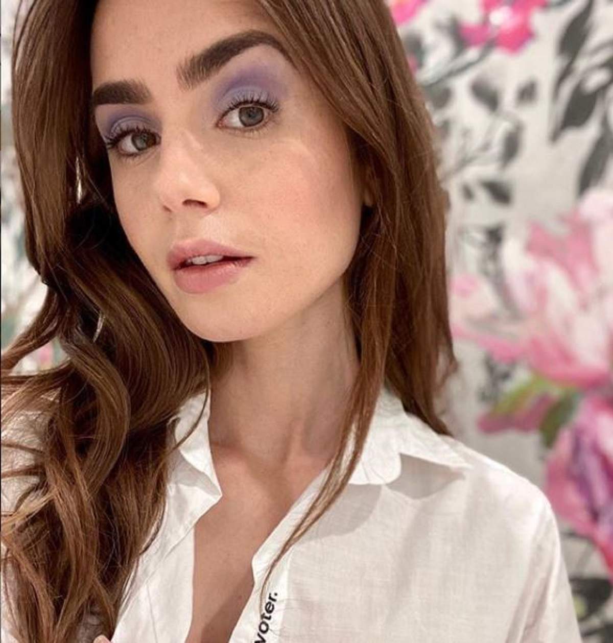 Lily Collins poarta o camasa alba si are in spate un panou cu flori