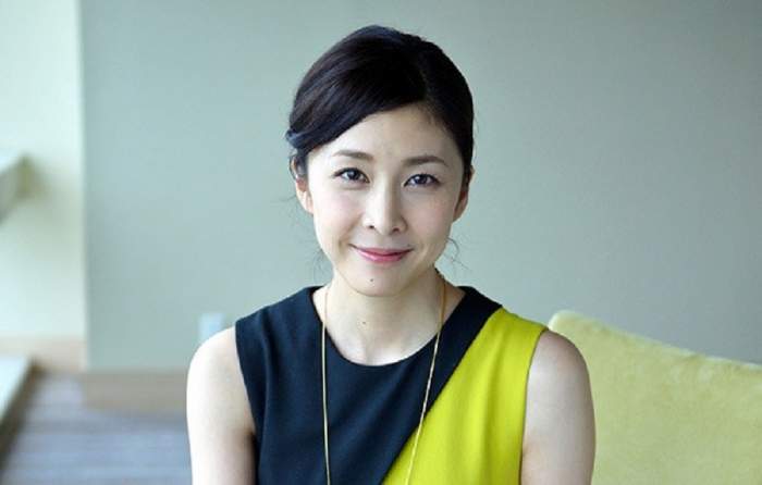Actrita Yuko Takeuchi sta pe o canapea, zambeste si poarta un top fara maneci, negru cu galben
