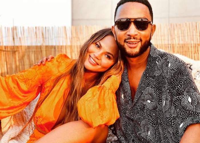 Chrissy Teigen și Jogn Legend se țin în brațe. Modelul poartă o rochie oranj, iar artistul o cămașă neagră cu picățele albe.