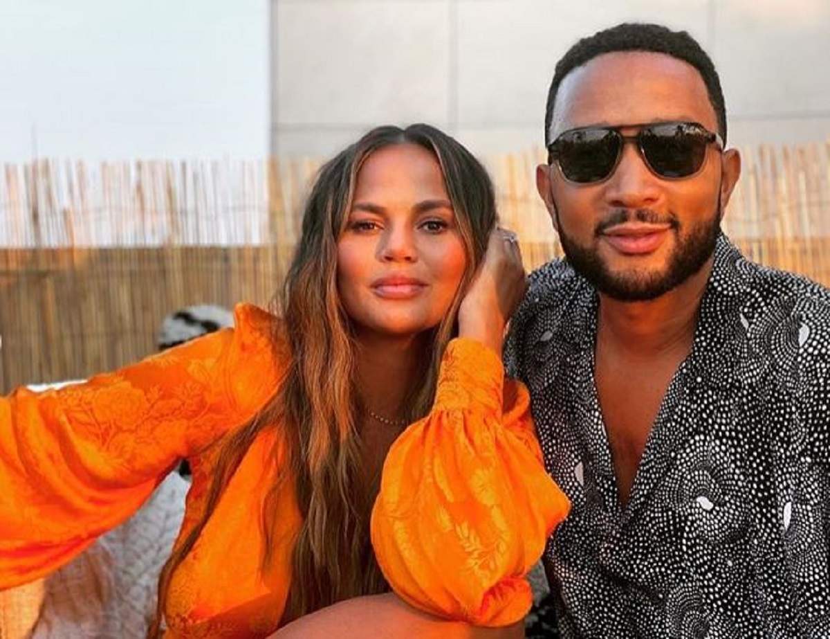 Chrissy Teigen și Jogn Legend se țin în brațe. Modelul poartă o rochie oranj, iar artistul o cămașă neagră cu picățele albe.