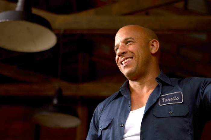 Vin Diesel poarta o camasa descheiata albastra si un tricou alb, Zambeste larg si se uita n fata