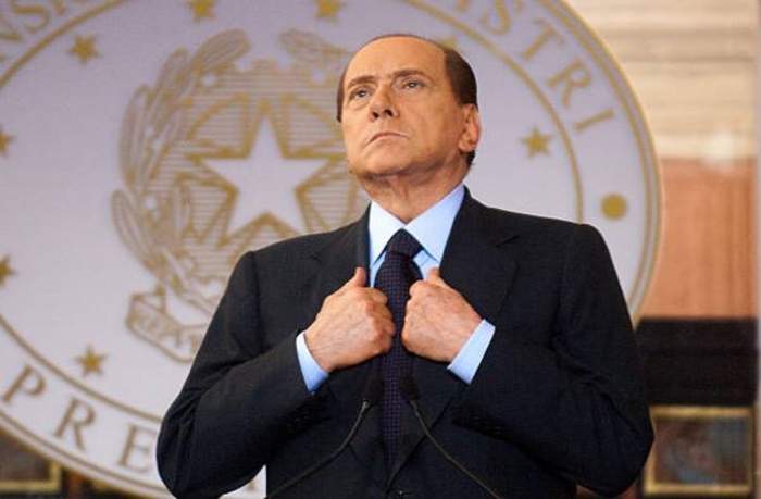 Silvio Berlusconi în Guvern, poartă un costum negru si pozează impunător în timp ce privește în sus