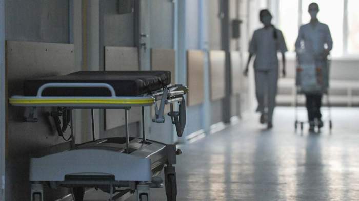 Două asistente medicale se plimbă pe holurile spitalului, cu un pat în prim plan
