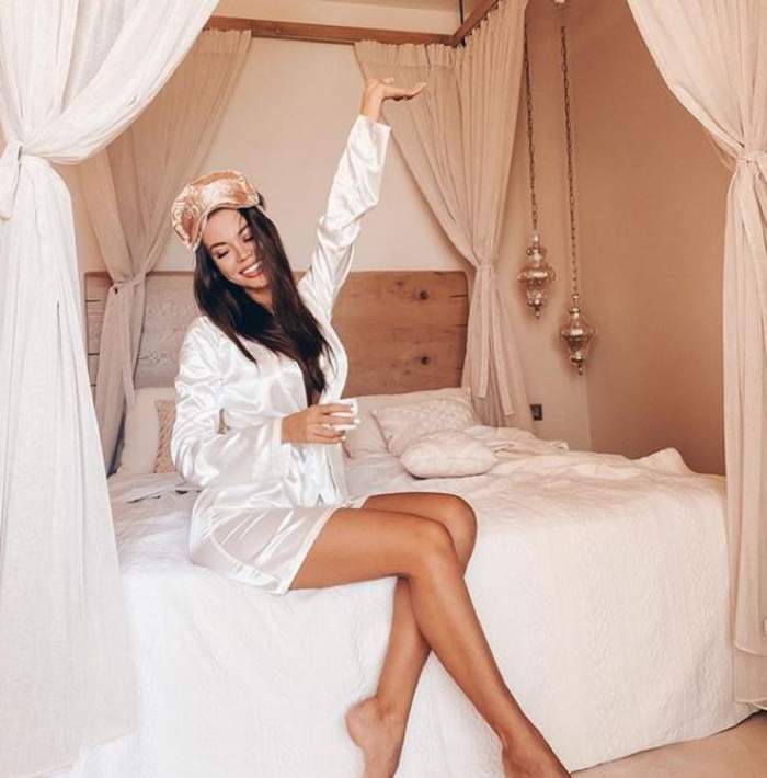 Denisa Hodișan este îmbrăcată cu un halat de culoare albă, în timp ce stă pe marginea patului, râde și stă cu o mână ridicată