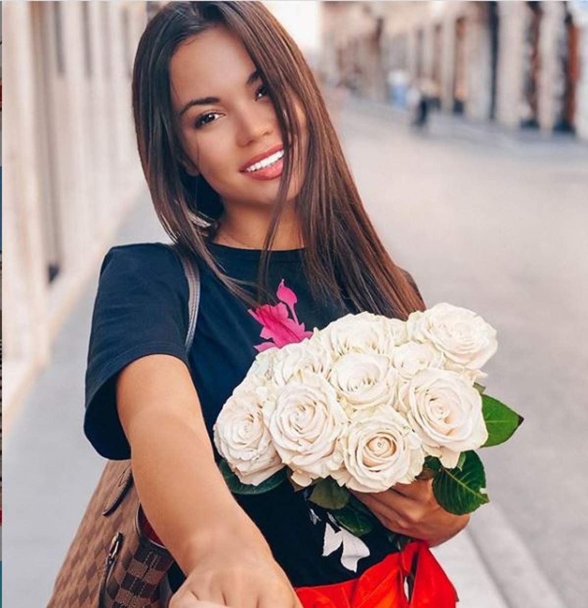 Denisa Hodișan ține un buchet de trandafiri albi în brațe, zâmbește la cameră și-și ține iubitul de mână