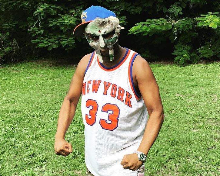 Rapperul american MF Doom se află afară. Artistul poartă un maiou alb și niște pantaloni camuflaj. Cântărețul are pe cap o șapcă albastră, iar pe față o mască.