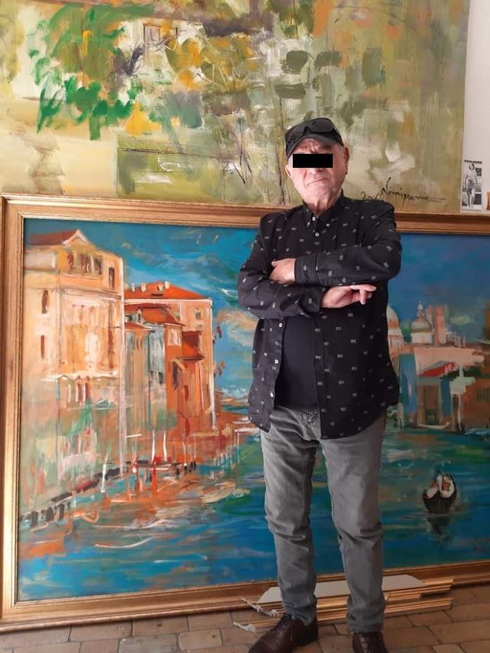 Viorel Nimigeanu purta o pereche de pantaloni gri, o cămașă neagră cu model alb și o șapcă neagră. Pictorul ținea mâinile în sân și lângă el era un tablou.