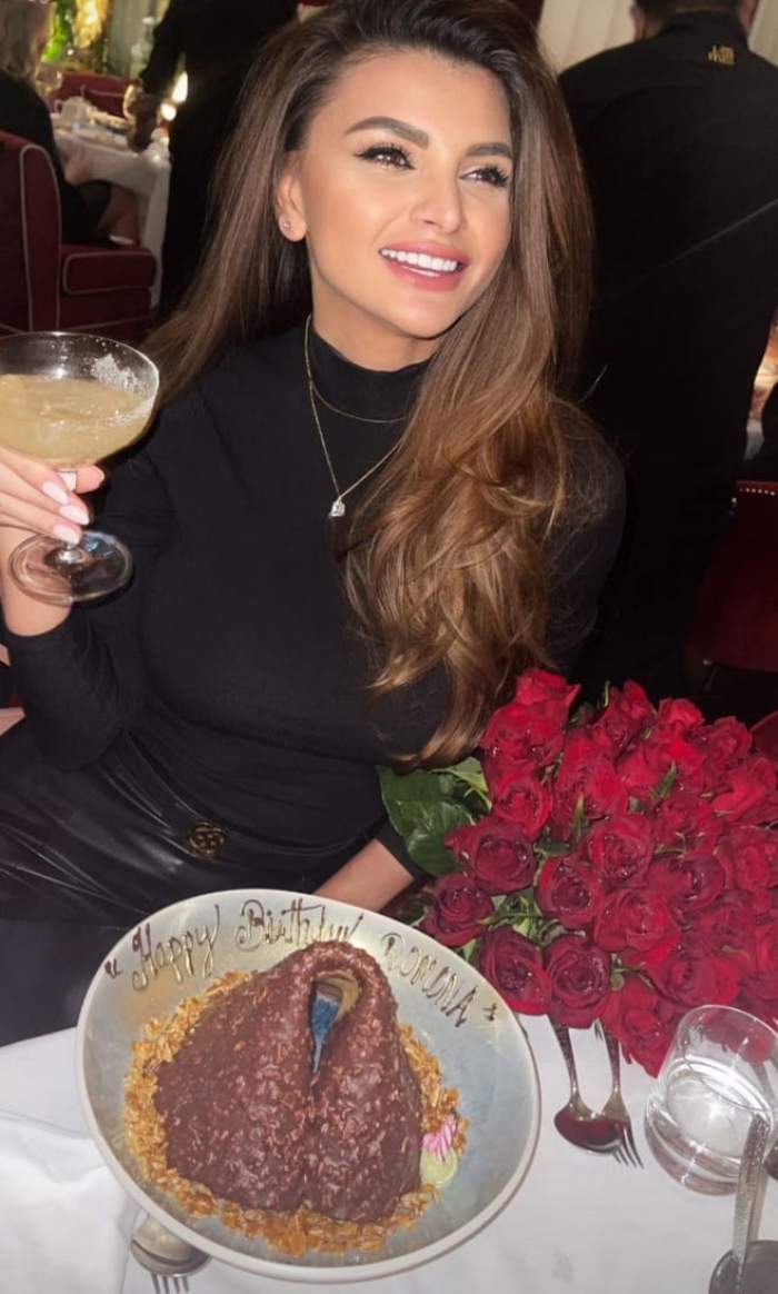 Ramona Gabor, la masă,  cu paharul în mână, cu tort în față și un buchet de trandafiri roșii alături
