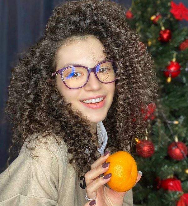 Cleopatra Stratan ține în mână o portocală. Artista e îmbrăcată în hanorac crem, poartă ochelari de vedere, iar în spatele ei se vede bradul de Crăciun.
