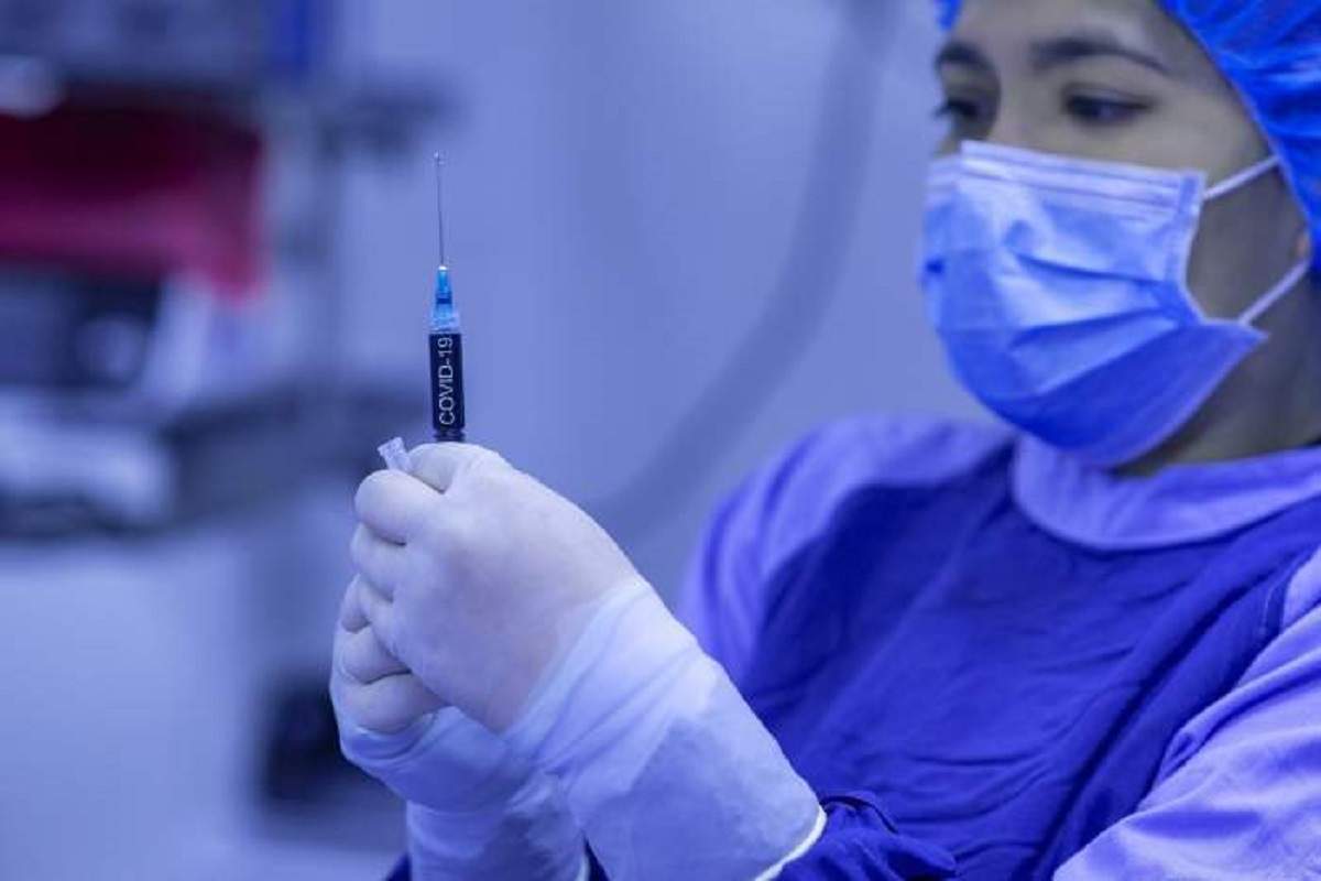 Un cadru medical ține în mână un vaccin anti-covid-19. Femeia poartă echipament special.