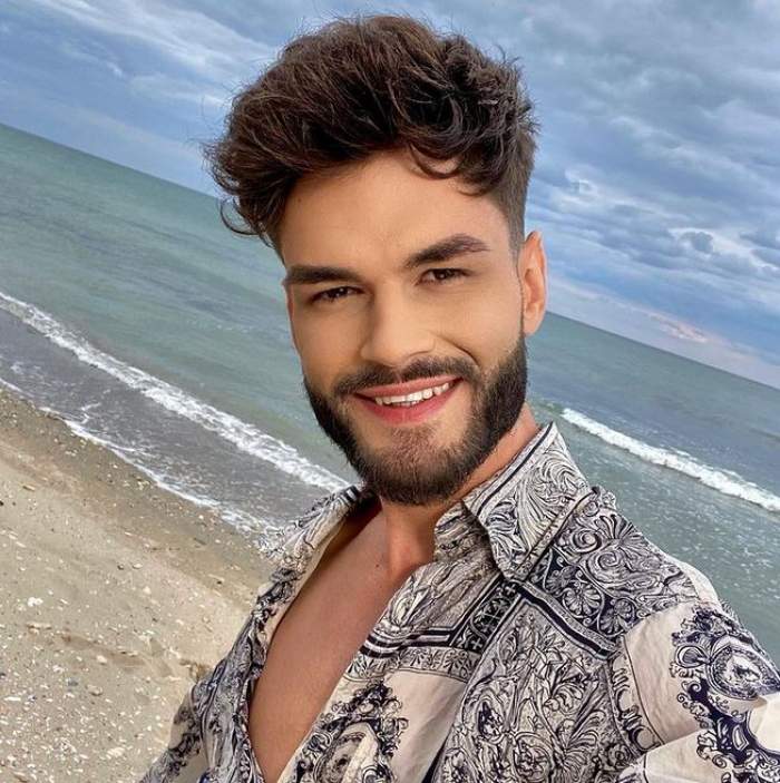 Edward Sanda este pe plajă. Artistul zâmbește larg și e îmbrăcat într-o cămașă albă cu detalii florale negre.