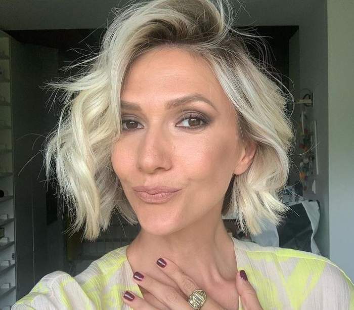 Adela Popescu își face un selfie. Vedeta poartă o bluză albă cu model galben și își ține mâna pe piept.