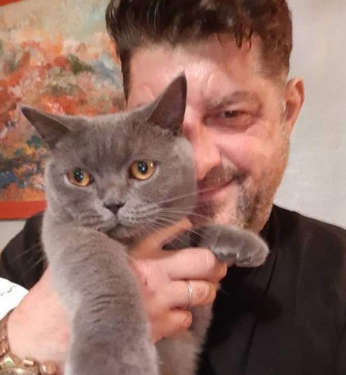 Tavi Colen poartă un tricou negru. Artistul o ține într-o mână pe pisica lui de culoare gri.