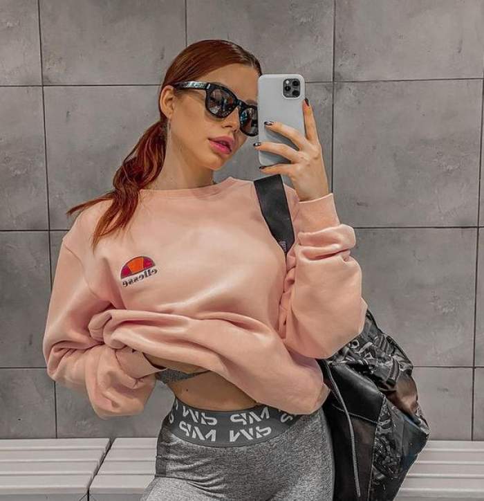 Carmen Grebenișan își face o poză în oglindă cu telefonul mobil. Vedeta poartă o bluză roz, niște colanți gri, iar pe umăr are o geantă neagră.